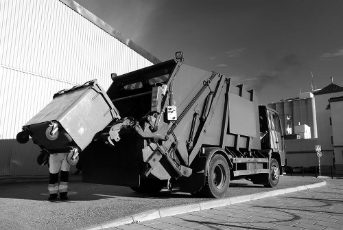 A black and white photograph showing a commercial rubbish collection vehicle parked on an urban street near a large corrugated metal warehouse wall. The vehicle has a hydraulic lift mechanism at the rear, used for lifting and emptying waste bins, which are visible attached to the truck. A worker in high-visibility clothing stands beside the vehicle, operating or inspecting the loading process. The waste bin, made of durable plastic with a textured surface, is being lifted into the truck's compartment. The scene is set on a paved roadway adjacent to a sidewalk, with other industrial buildings faintly visible in the background under a partly cloudy sky. This image captures the routine activity of rubbish collection associated with local rubbish removal services like Office Clearance Kenton, serving the nearby Kenton area and postcode.