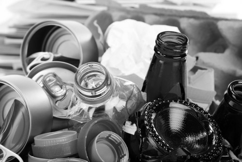 A collection of discarded household waste materials including glass bottles, metal cans, plastic containers, and lids, piled together on a surface in an outdoor area likely near a property in Kenton. The glass bottles are transparent with some appearing empty, with black and white reflective surfaces, while the plastic containers and lids vary in size and shape, primarily in shades of grey. In the background, there are some crumpled paper or tissue items, suggesting general rubbish. The environment appears to be part of a rubbish removal scene, typical of a Service provided by Office Clearance Kenton, with the waste materials arranged loosely and yet ready for collection or disposal. The overall scene indicates a typical cluttered outdoor space prepared for rubbish clearance, with the objects occupying a significant portion of the foreground and middle ground, set in natural lighting that highlights textures and the diverse materials. This detailed description supports understanding of rubbish removal processes in a London suburb context, such as Kenton in the borough of Brent, where local services often handle similar waste collection tasks.