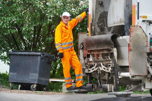 A waste collection operative dressed in high-visibility orange work clothing and a white cap standing next to a large industrial rubbish compactor on a paved area with green leafy trees in the background. The worker is positioned on the right side of the image, slightly leaning against the compactor, which has a large metal roller and a side panel with safety markings. To the left, a black wheeled rubbish bin is placed on the pavement, ready for waste collection. The scene appears to be outdoors in a residential or commercial area, with natural light illuminating the scene. The image relates to rubbish removal services provided by Office Clearance Kenton, emphasizing efficient waste management and recycling efforts relevant to the local area.