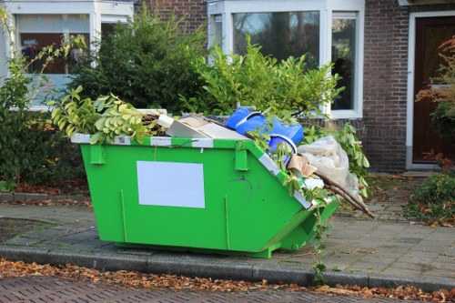 A large green skip filled with assorted rubbish, including green and white cardboard boxes, plastic packaging, and some plant material, positioned on a pavement outside a residential building in Kenton. The skip is situated next to a concrete curb and surrounded by green shrubbery and tree branches. In the background, there are windows and a door of a brick house, with a garden area visible. The waste appears to be a mixture of household items and garden debris, typical of the clearance services provided by Office Clearance Kenton for domestic or small-scale commercial rubbish removal in the area.