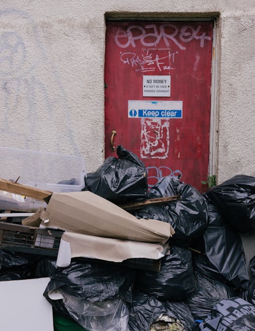 A large pile of assorted metal debris and broken building materials is situated outside a partially constructed or abandoned multi-storey building. The rubble includes twisted metal beams, shattered p