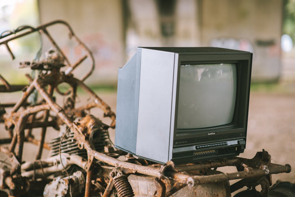 A collection of discarded electronic appliances and waste items is displayed outdoors on a sandy surface, consisting mainly of old CRT televisions with large, boxy screens, many of which have visible