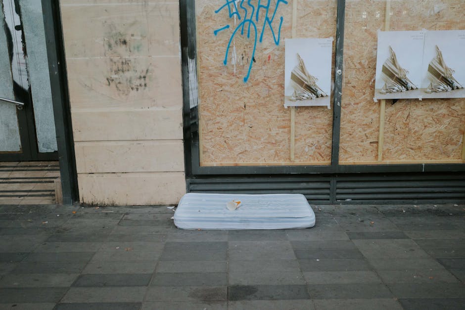 A person wearing a black jacket and a patterned headband is standing outdoors on a city street corner, carrying a large, padded, black and green mattress with orange handles. The mattress appears to b