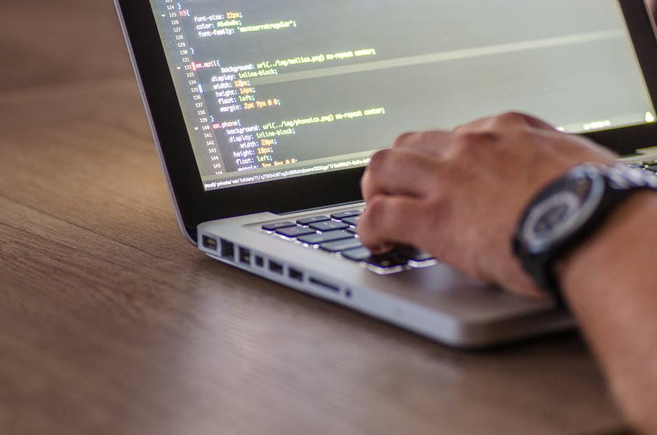 A person's right hand, wearing a wristwatch with a black strap and round face, operates a laptop with a silver body and a black keyboard on a wooden surface. The laptop screen displays lines of coding