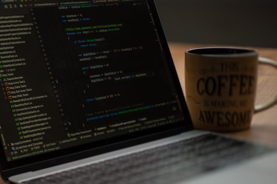 A close-up image of a laptop placed on a wooden desk, displaying lines of colourful programming code on its dark screen. To the right of the laptop, there is a coffee mug with a beige exterior featuri