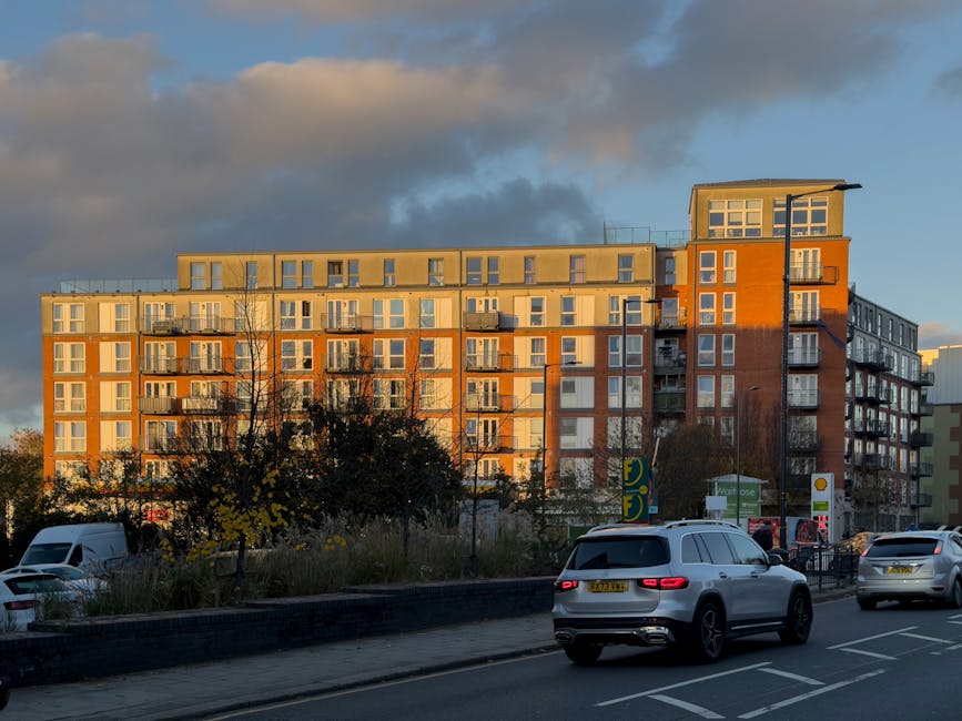 A red passenger van parked on a city street in front of a three-story residential building with a brick facade featuring alternating yellow and red panels. The van is positioned parallel to the paveme